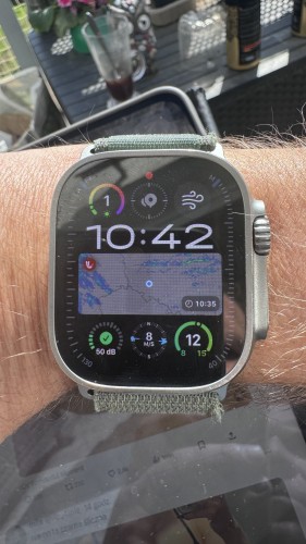 Smartwatch na nadgarstku wyświetlający różne widżety, w tym godzinę, pogodę, poziomy dźwięku i pierścienie aktywności. Tło wydaje się być scenerią zewnętrzną ze szklanką i przedmiotami na stole.