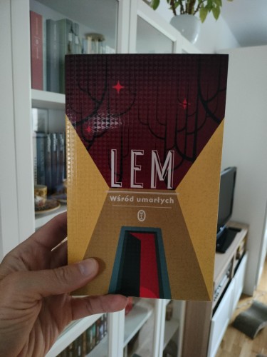 Zdjęcie przedstawia książkę trzymaną w lewej ręce. Okładka ma nowoczesny, minimalistyczny design w odcieniach żółci, czerwieni i ciemnego bordo. Na górze widnieje nazwisko autora: "LEM", a poniżej tytuł książki: "Wśród umarłych". W tle grafiki widać stylizowane, czarne zarysy drzew i czerwone gwiazdy, a centralnie umieszczona jest abstrakcyjna forma przypominająca otwarte drzwi. W tle zdjęcia znajduje się biały regał z książkami, szklana witryna, a na górze widać zwisającą doniczkę z rośliną.