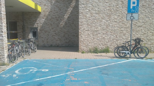 Empty bicycle rack in the middle of the photo. The bicycle rack is of the "lock up only the wheel to make it easier to steal the rest of the bike" variety.
Around it there are four bicycles: two chained to the shopping carts corral, and two to the parking sign. 