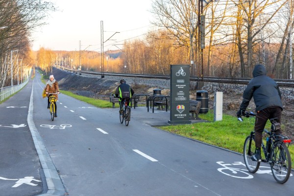 zdjęcie velostrady, na niej troje rowerzystów, po prawej stronie od velostrady leci linia kolejowa, po lewej umiejscowiono chodnik, w tle widać miejsca postojowe z ławkami i koszami na smieci