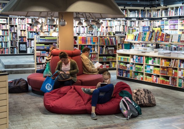 Zdjęcie z księgarni lub biblioteki, gdzie wokół jest pełno półek z kolorowymi książkami, a na środku stoją pufy i miękkie fotele. Siedzą na nich ludzie (w tym jedno dziecko), wszyscy pogrążeni w lekturze. Nad nimi jest masa lampek. Źródło zdjęcia: https://www.pexels.com/pl-pl/zdjecie/pelna-dlugosc-mezczyzny-siedzacego-na-podlodze-256431/