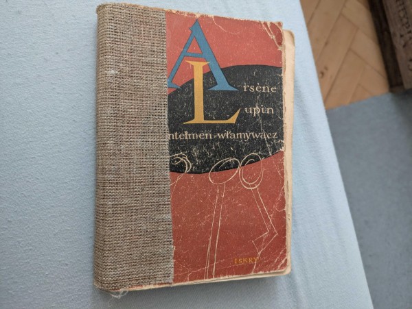 Zdjęcie zniszczonej okładki książki "Arsen Lupin. Dżentelmen Włamywacz" autorstwa Maurice'a Leblanca. Okładka jest w odcieniach ciemnej czerwieni i widać, że to stara pozycja.