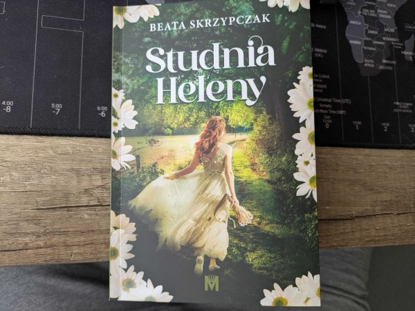 Zdjęcie książki "Studnia Heleny" Beaty Skrzypczak. Okładka ukazuje kobietę w białej sukni z bukietem kwiatów, która przechadza się lub biegnie pomiędzy drzewami, po trawie blisko jeziora.
