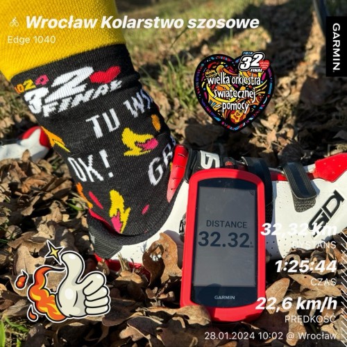 Stopa rowerzysty w ozdobnej skarpecie obok komputera rowerowego Garmin wyświetlającego dane jazdy, z naklejkami i emblematami związanymi z 32 finałem WOŚP. Na ziemi rozrzucone są liście.