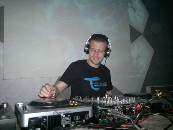Ja, w okularach i słuchawkach siedzę za konsoletą DJ-ską marki Pioneer, z wyraźnym uśmiechem na twarzy. W tle klubowe otoczenie, z projekcją świetlną w tle.