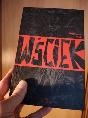 Dłoń trzymająca książkę Wściek Magdaleny Salik. Ciemna okładka z ilustracją gęstego, mrocznego lasu; przez środek przebiega duży czerwony tytuł »WŚCIEK«. Na dole widoczne logo wydawnictwa Powergraph.