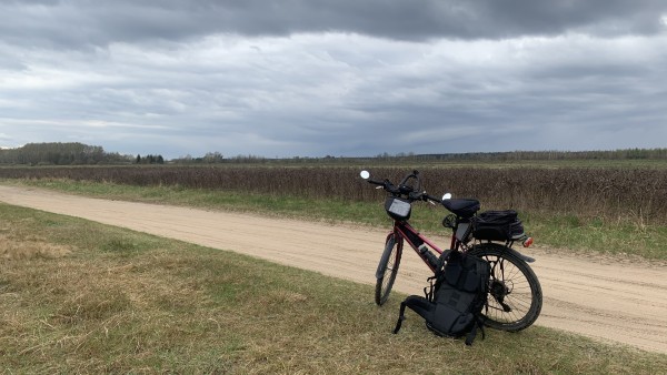 Rower retro mtb na trawie przed piaszczystą drogą polną. W tle niska uprawa nie wiem czego. Dalej las, część nieba zachmurzona, część na górze zdjęcia bardzo ciemna.