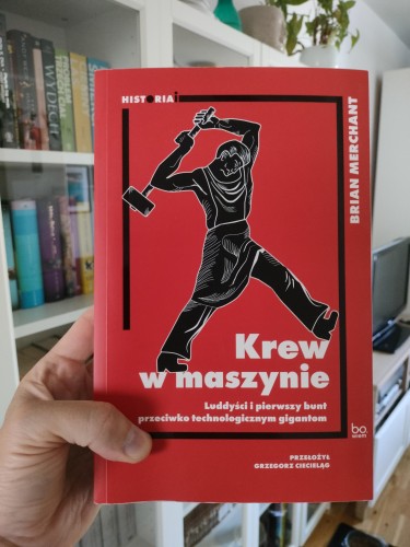 Okładka książki trzymanej w dłoni na tle domowego regału z książkami. Na czerwonym tle widnieje grafika w stylu drzeworytu przedstawiająca człowieka zamachującego się młotem. Tytuł książki: Krew w maszynie. Luddyscy i pierwszy bunt przeciwko technologicznym gigantom. Autor: Brian Merchant. Przekład: Grzegorz Cieciełag. Wydawnictwo: Bo.wiem.
