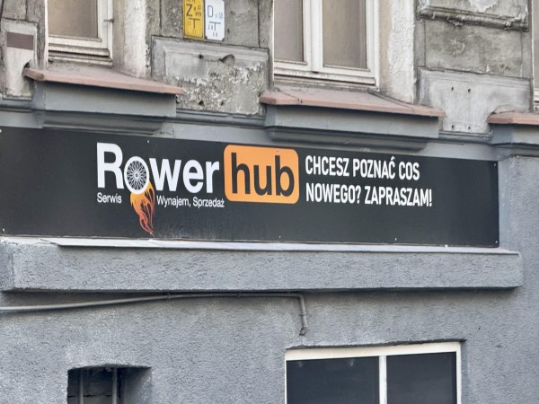 Znak dla “Rower Hub”, z grafiką koła rowerowego, promujący usługi związane z rowerami, w tym wynajem i sprzedaż. Zawiera tekst w języku polskim zachęcający ludzi do poznania czegoś nowego.