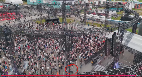 Widok z góry na duży festiwal muzyczny na otwartym terenie. Tłum ludzi gromadzi się przed sceną, otoczoną masywną, czarną konstrukcją z kratownic stalowych, na których zamocowane są światła i nagłośnienie. W tle widoczne są namioty i stoiska, a w dolnej części zdjęcia czerwonym kółkiem zaznaczono osobę na wózku.