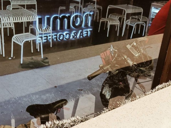 Photo of a bike parked outside, shot through the glass window from inside a cafe. There is a phone and a light mounted on the handlebar. There is also a solar powerbank and a helmet hanging from the handlebar.