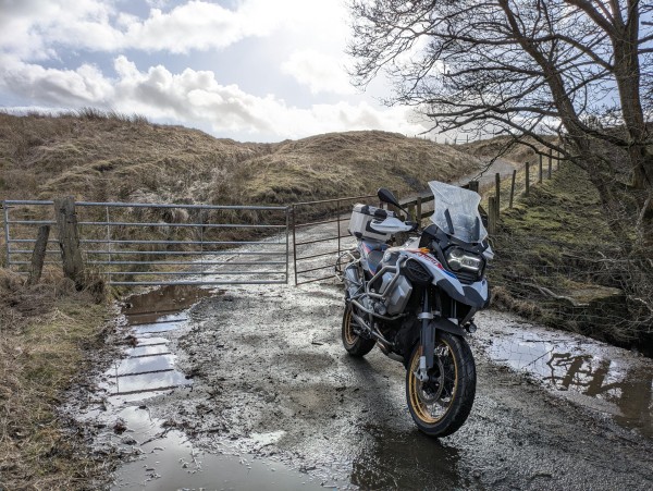 A wet and muddy single track road. There is a gate across the road. A motorcycle is parked in front of the gate, waiting to go.