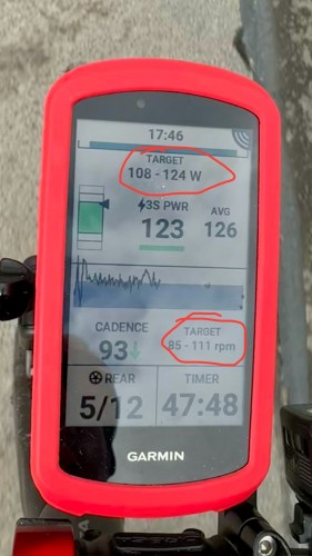 Komputer rowerowy Garmin wyświetlający różne wskaźniki jazdy, takie jak moc, kadencja i czas, zamontowany na rowerze. Ekran pokazuje docelowe strefy mocy i kadencji, zakreślone na czerwono.