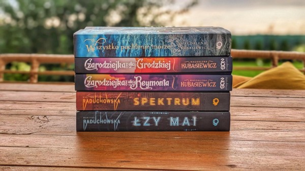 Na drewnianym stole leży stos pięciu używanych książek. Trzy z nich są autorstwa Magdaleny Kubasiewicz i dwie Martyny Raduchowskiej. Grzbiety książek są widoczne — od góry: „Wszystko pochłonie morze” (Magdalena Kubasiewicz, wydawnictwo SQN), „Czarodziejka z ulicy Grodzkiej”, „Czarodziejka z ulicy Reymonta” (obie Magdalena Kubasiewicz, SQN), „Spektrum” (Martyna Raduchowska) oraz „Łzy Mai” (Martyna Raduchowska). W tle widać rozmytą, zieloną scenerię i elementy drewnianej ławki z poduszką, co sugeruje, że zdjęcie wykonano na tarasie lub werandzie. Książki wyglądają na dobrze zachowane, lecz używane.
