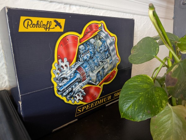 A box of a Rohloff speedhub, a bicycle part, with a green plant in front of it.