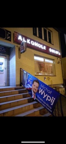 Plakat wyborczy Michała Wypija na poręczy schodów do sklepu „Alkohole Świata”
