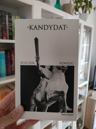 Zdjęcie przedstawia książkę trzymaną w dłoni na tle domowego regału z książkami. Okładka książki jest utrzymana w minimalistycznym stylu, z tytułem „Kandydat” u góry, nazwiskiem autora „Żulczyk” po lewej i określeniem „Powieść” po prawej. Centralną część okładki zajmuje czarno-białe zdjęcie pomnika jeźdźca z uniesionym mieczem. W prawym dolnym rogu znajduje się logo wydawnictwa „Świat Książki”.