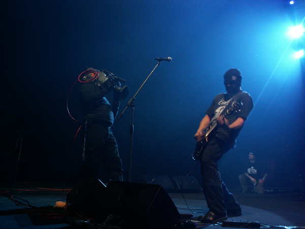Scena w "Spodku", koncert metalowy, po lewej stronie kamerzysta, po prawej - gitarzysta, obaj ubrani na czarno, zabawnie odgięci, jakby w tańcu.