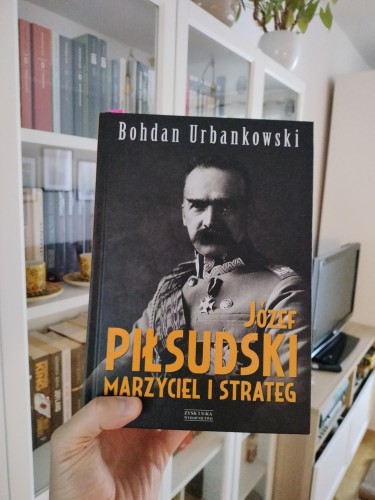 Na zdjęciu widzimy osobę trzymającą książkę Bohdana Urbankowskiego zatytułowaną „Józef Piłsudski. Marzyciel i strateg”, wydaną przez Wydawnictwo Zysk i S-ka. Okładkę zdobi czarno-biała fotografia Józefa Piłsudskiego w mundurze, z charakterystycznym wąsem, na ciemnym tle. Tło zdjęcia to przytulne wnętrze mieszkania – widać półki z książkami, filiżanki oraz telewizor i rośliny doniczkowe.