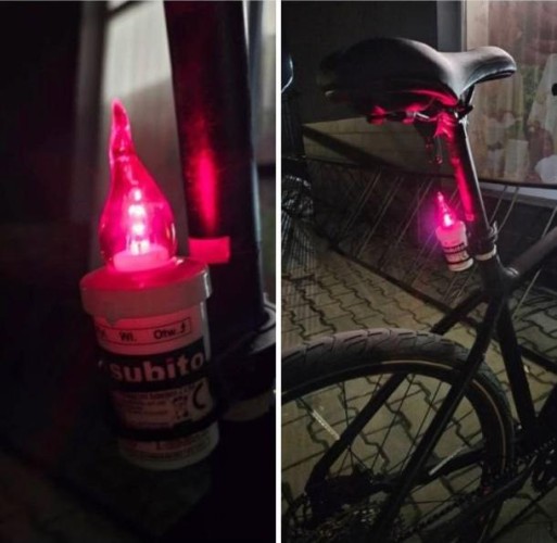 Na zdjęciu widzimy nietypową tylną lampkę rowerową. Jest to czerwone, plastikowe światło przypominające znicz nagrobny, zamontowane na wsporniku siodła roweru. Lampka emituje czerwoną poświatę i została przymocowana do ramy roweru przy pomocy opaski zaciskowej. Tło to nocne otoczenie, a rower stoi zaparkowany na chodniku z kostki brukowej, w pobliżu przeszklonej ściany budynku.