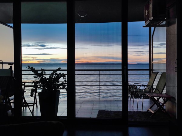 Widok z pokoju / apartamentu na jezioro Balaton, wieczór, chmury i niebo w kolorach niebieskim, granatowym, pomarańczowym, na pierwszym planie widoczny balkon z krzesłami, stolikiem i roślinką w dużej donicy
