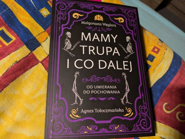 Okładka książki "Mamy trupa i co dalej. Od umierania do pochowania" autorstwa Małgorzaty Węglarz i Agnes Tołoczmańskiej. Książka w fioletowo-czarnej stylistyce, ze szkieletami i zawijasami.