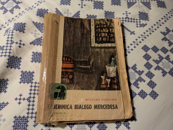 Poklejona okładka książki "Tajemnica białego mercedesa" Nicolasa Freelinga. Książka jest z serii z Jamnikiem, pokazuje pokój z winem i kieliszkami na biurku, a także fotel.
