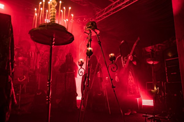 Słabo oświetlona scena z czerwonym oświetleniem przedstawiająca muzyków w szatach występujących wśród kilku świeczników i atmosferycznej mgły.