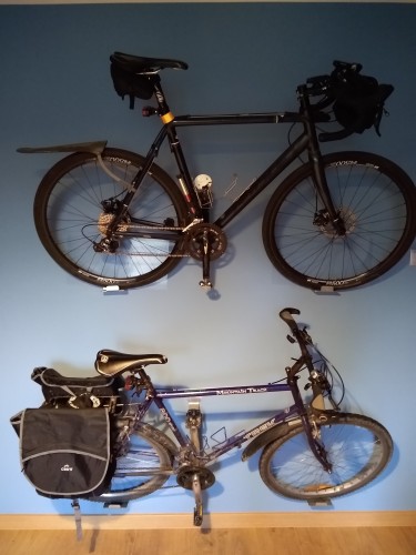 Dwa rowery wiszą na niebieskiej ścianie. Wyżej wisi szutrówka, a poniżej rower trekingowy z sakwami na bagażniku.