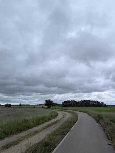 Wiejska ścieżka, a obok niej ścieżka rowerowa. Obie otoczone polami i skręcające w lewo. W tle widać jadących rowerzystów oraz drzewa. Pochmurne niebo.