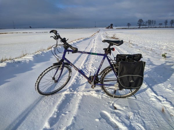 Rower z sakwami na bagażniku stoi w poprzek polnej drogi zasypanej śniegiem.