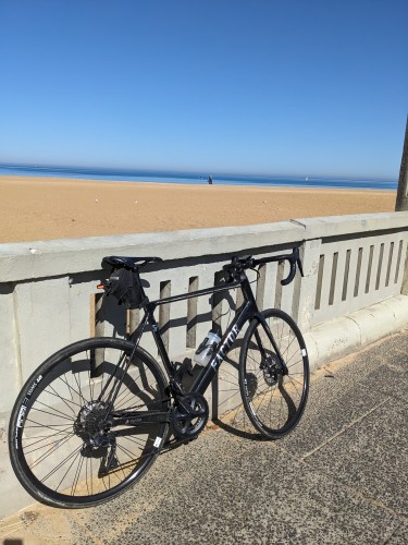 A black bike leaning against a concrete fence seperating a path from a beach.  On the far side of the fence is an unpopulated sandy beach, with still water and a glorious blue sky.