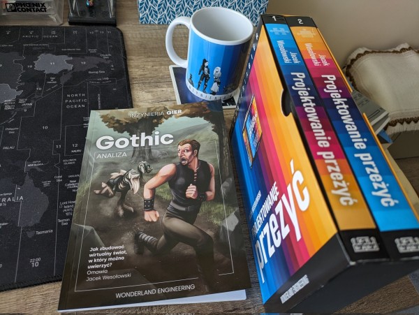 Na biurku widać dwa tomy "Projektowania przeżyć" Jacka Wesołowskiego, książeczkę "Gothic - analiza" tego samego autora oraz kubek "Inżynieria gier" w barwach tęczy z postaciami.