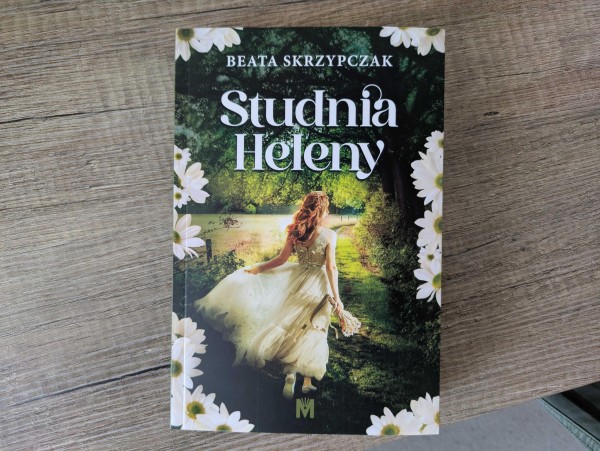 Okładka książki "Studnia Heleny" Beaty Skrzypczak. Okładka ukazuje kobietę w białej sukni z bukietem kwiatów, która przechadza się lub biegnie pomiędzy drzewami, po trawie blisko jeziora.