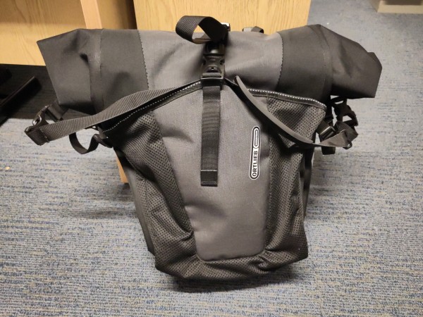 One of a pair of bike panniers, pictured as you'd see it from the side of the bike. The roll top is secured with one clip, and rolls back towards the rack when it's on the bike.

It also has a shoulder carrying strap, and a big, partly mesh, side pocket. The pocket is secured with its own clip, which I also use to secure the carry strap when in motion. Dead easy to do before setting off.

It has some holders for securing the strap somewhere underneath too, which you're supposed to use, but this is much more convenient.