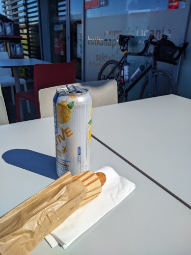 Na stoliku przy stacji paliwowej leży hotdog, a obok stoi puszka piwa. W tle widać rower, oparty o ścianę stacji.